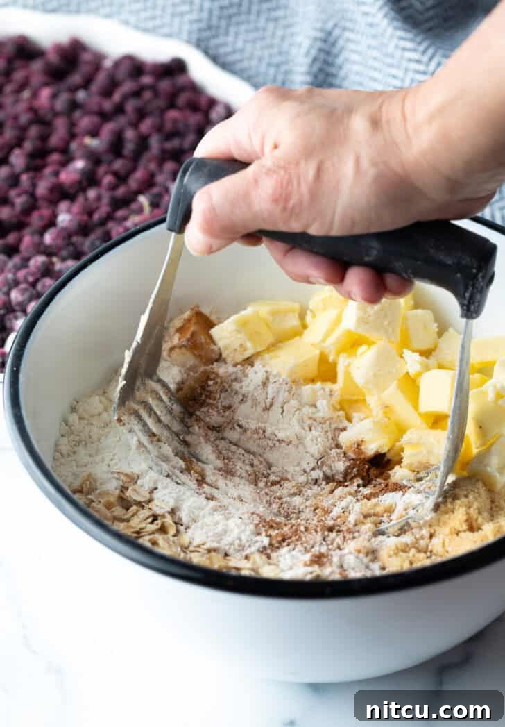 Perfect Blueberry Crisp 7 A close-up shot of a hand using a metal pastry cutter to cut cold butter into a mixture of flour, oats, and brown sugar in a bowl, creating a crumbly topping.