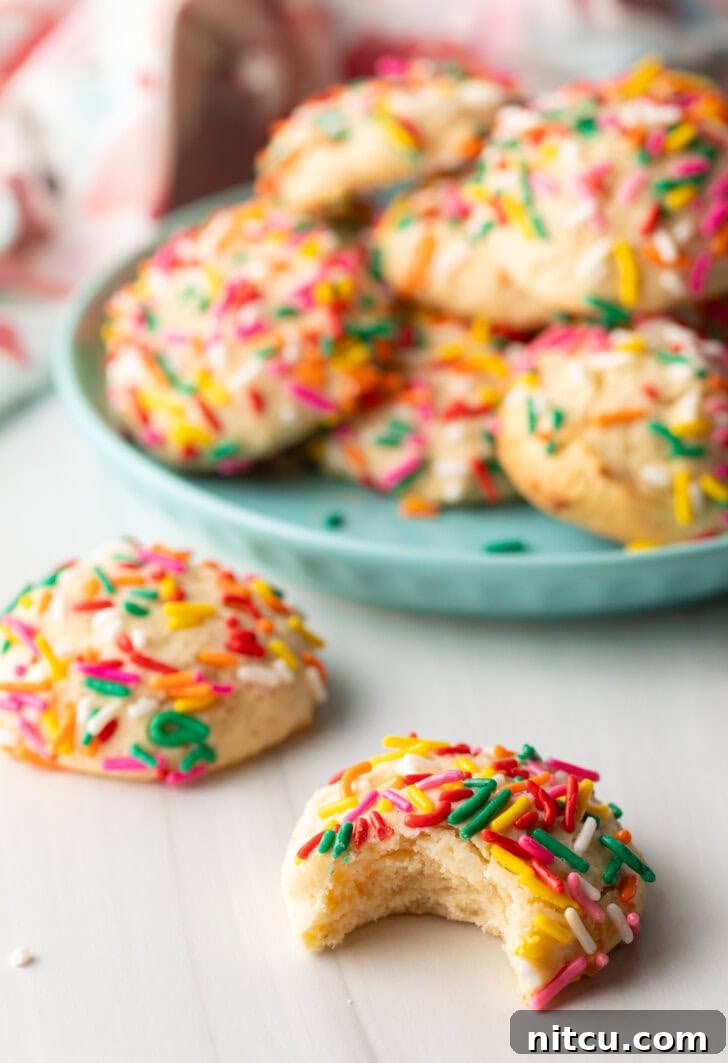 Velvety Cream Cheese Bites 4 Cookie with sprinkles and a bit taken out is close to camera. Another cookie with no bite is off to the side. In the background is a plate piled up with cream cheese cookies.