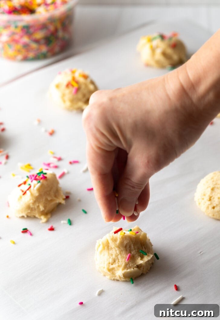 Velvety Cream Cheese Bites 6 Hand adding colorful sprinkles to the tops of raw cookies balls spread out on a baking sheet.