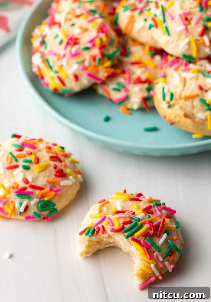Velvety Cream Cheese Bites 9 Cookie with sprinkles and a bit taken out is close to camera. Another cookie with no bite is off to the side. In the background is a plate piled up with cream cheese cookies.