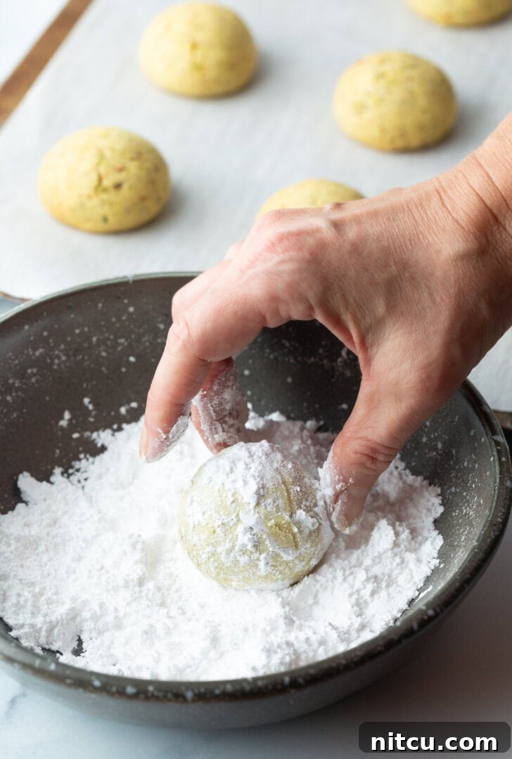 A hand gently rolling a warm, round cookie in a bowl filled with powdered sugar, ensuring full coverage.