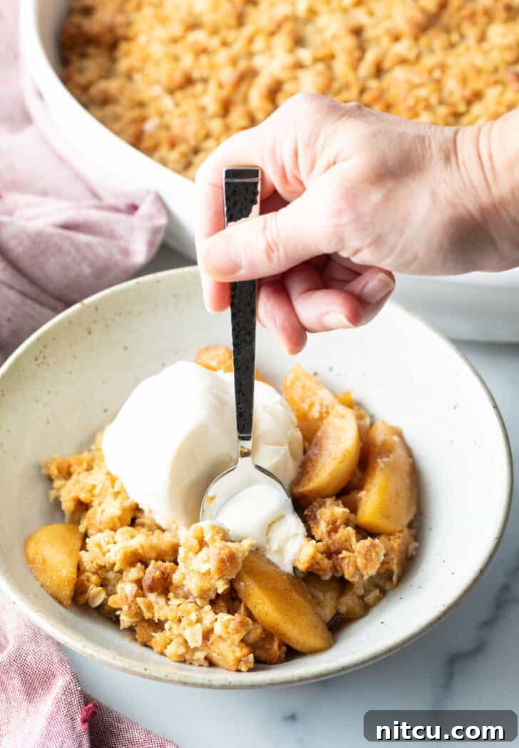Effortless Golden Apple Crisp 12 White bowl with golden apple crisp topped with a big scoop of ice cream. A hand with a metal spoon taking a bite of ice cream, showing pure enjoyment.