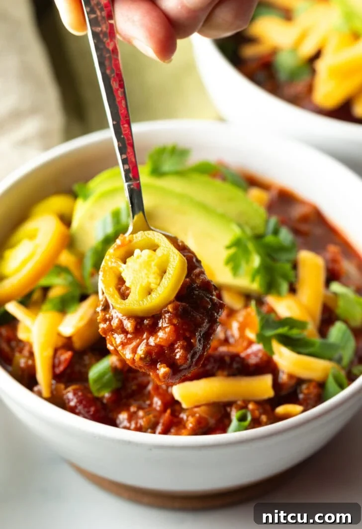 A steaming bowl of venison chili, generously topped with bright pickled jalapeno peppers, shredded cheese, creamy avocado slices, and finely chopped green onions, ready to be enjoyed.