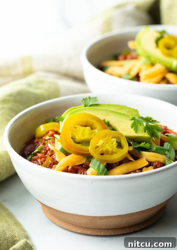 A beautifully composed bowl of venison chili, featuring pickled jalapeno peppers, a sprinkle of shredded cheese, fresh avocado slices, and chopped green onions as vibrant garnishes.