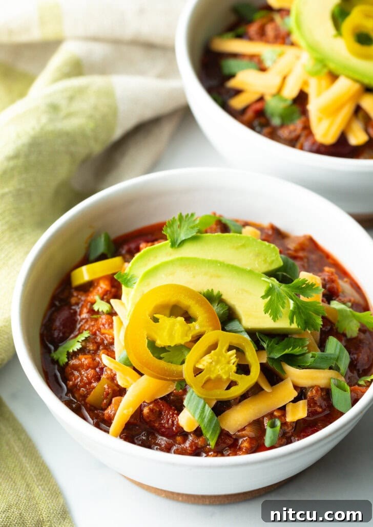 A final, appealing shot of the venison chili bowl, garnished perfectly with pickled jalapeno peppers, shredded cheese, avocado slices, and green onions, inviting you to take a bite.