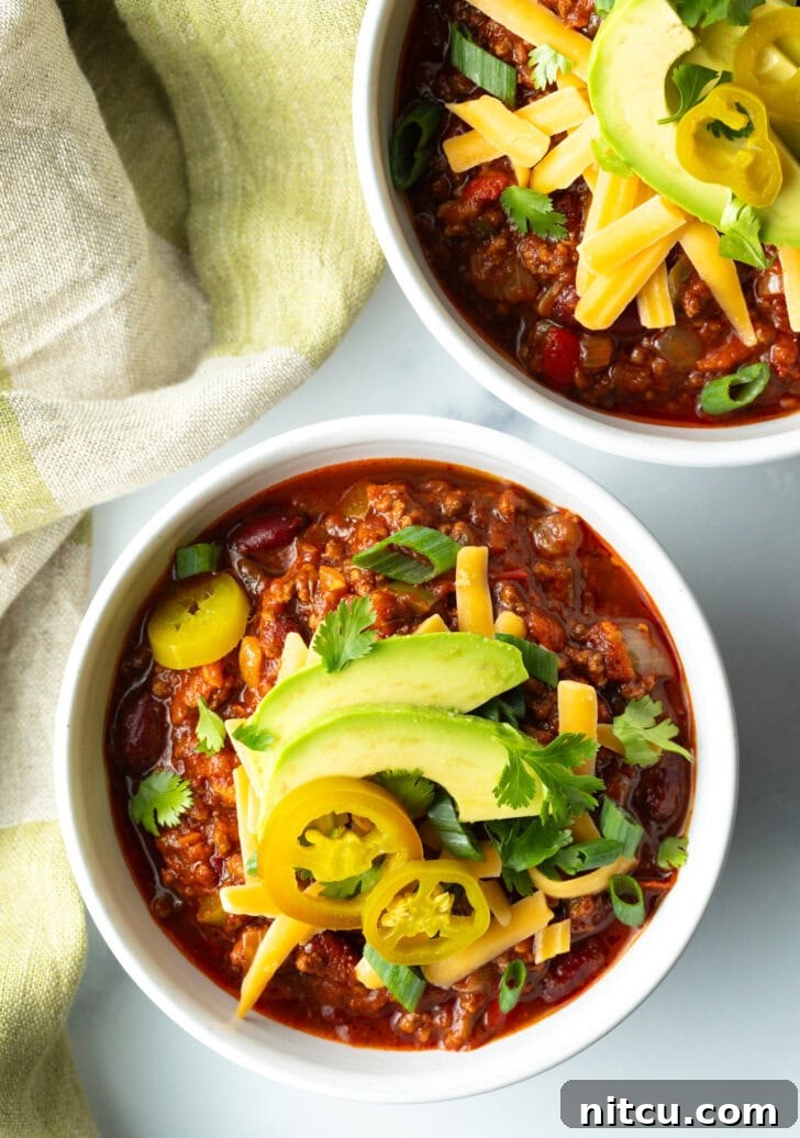 An overhead view of a rustic bowl filled with steaming venison chili, artfully garnished with vibrant pickled jalapeno peppers, a sprinkle of shredded cheese, fan-shaped avocado slices, and fresh green onions.