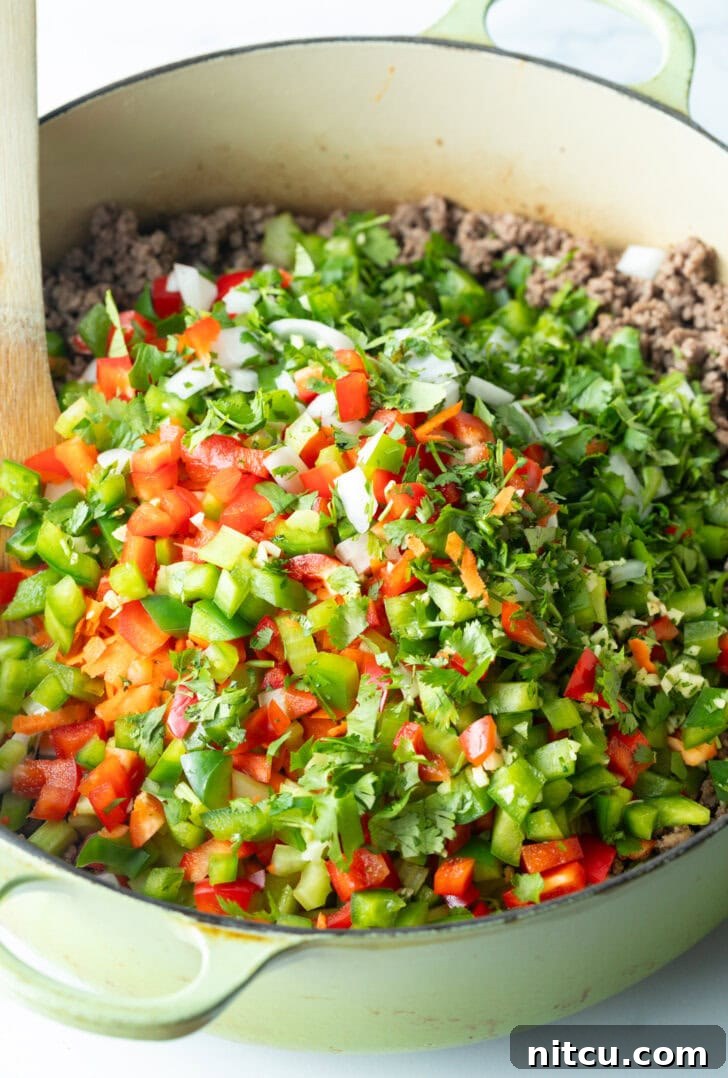 Adding freshly chopped bell peppers, onions, celery, and herbs to the pot with browned ground venison.