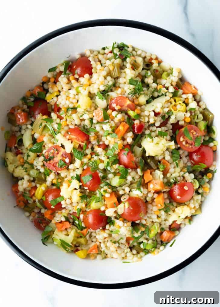 Top-down view of a colorful pearl couscous salad with chopped tomatoes, fresh herbs, and pickled vegetables in a large white mixing bowl, perfectly tossed.