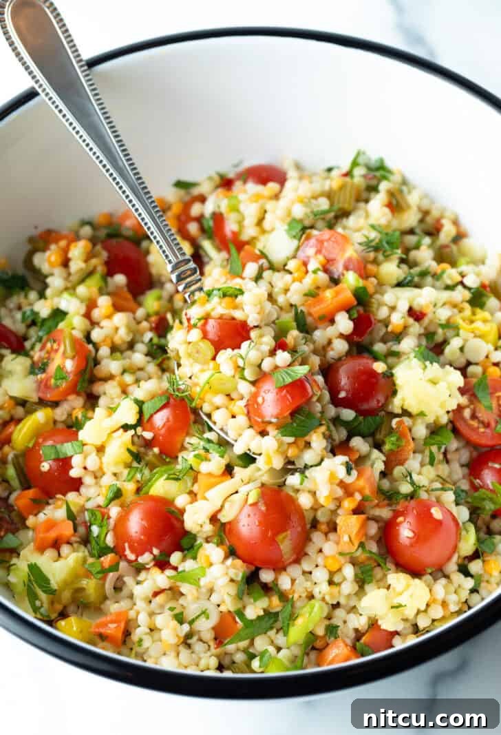 Pearl couscous salad with chopped tomatoes, fresh herbs, and pickled vegetables in a large white mixing bowl, perfectly prepared.