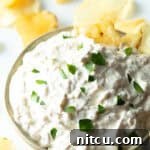 Bowl of creamy white French onion dip, topped with chopped chives and with a potato chip stuck into the dip.