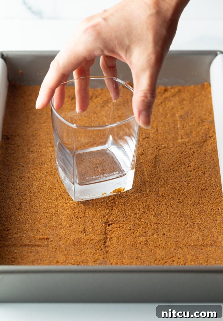 Sheet Pan No-Bake Pumpkin Cheesecake 7 Hand with glass pressing the cookie base into the bottom of a baking dish.