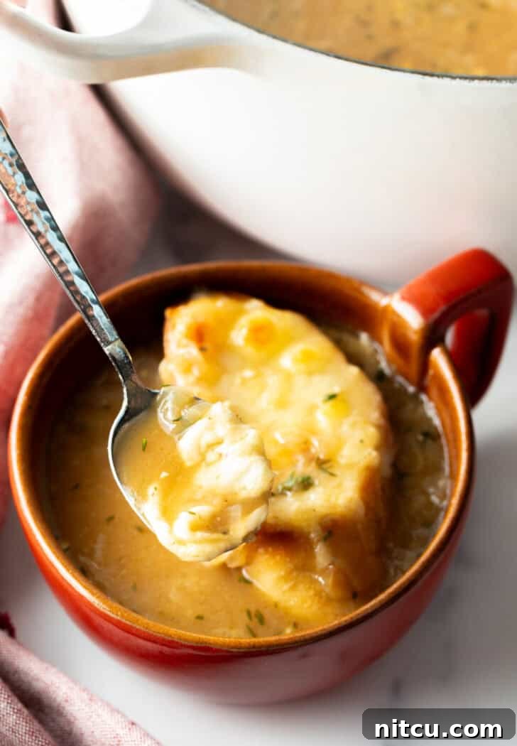 Bistro Onion Comfort 13 Large red mug with French onion soup, topped with a slice of baguette with melted cheese. A spoon is going in for a scoop.