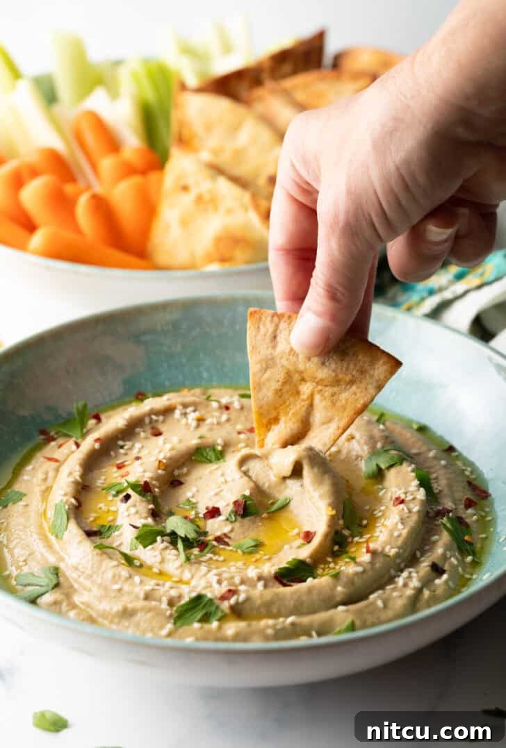 Hand using a pita chip to scoop creamy eggplant dip from a light blue bowl, garnished with a drizzle of olive oil.