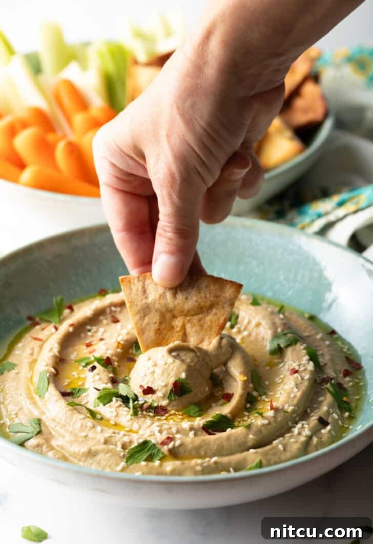 Hand using a pita chip to scoop eggplant dip from a light blue bowl, showcasing the creamy texture.
