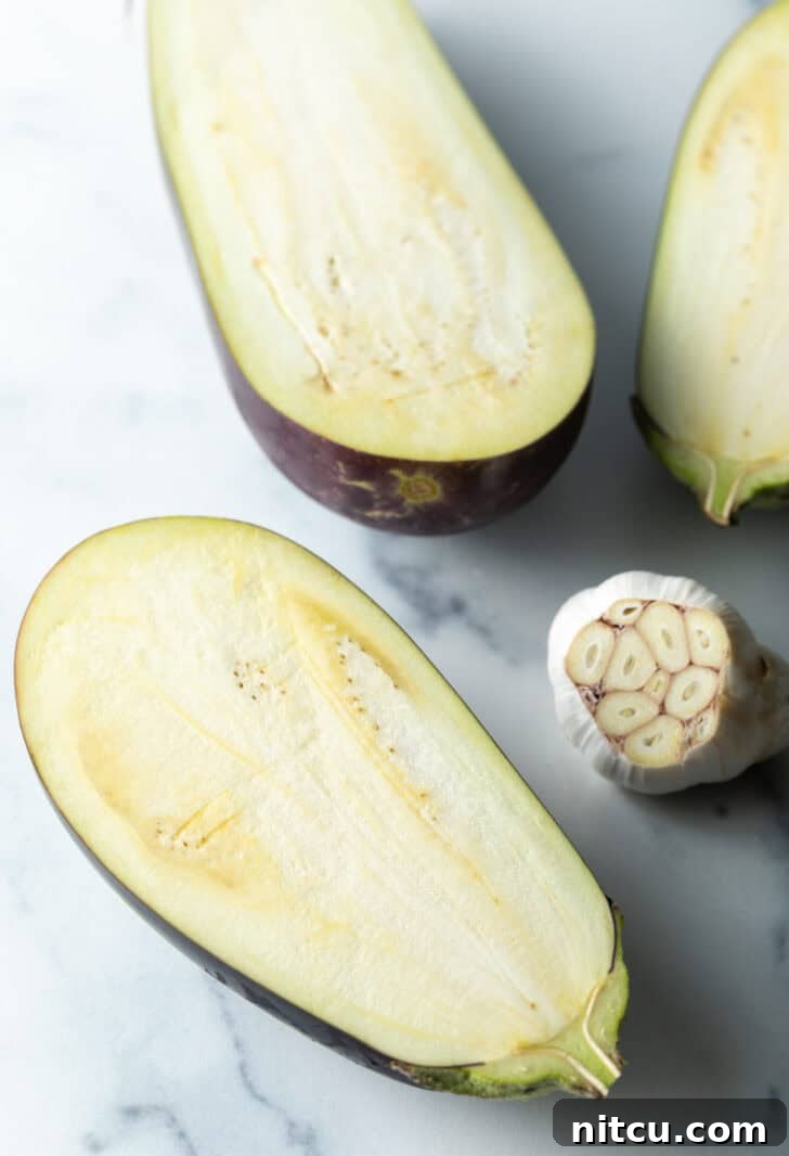 A raw eggplant halved, revealing its pale flesh and seeds, placed next to a head of garlic with the top chopped off.