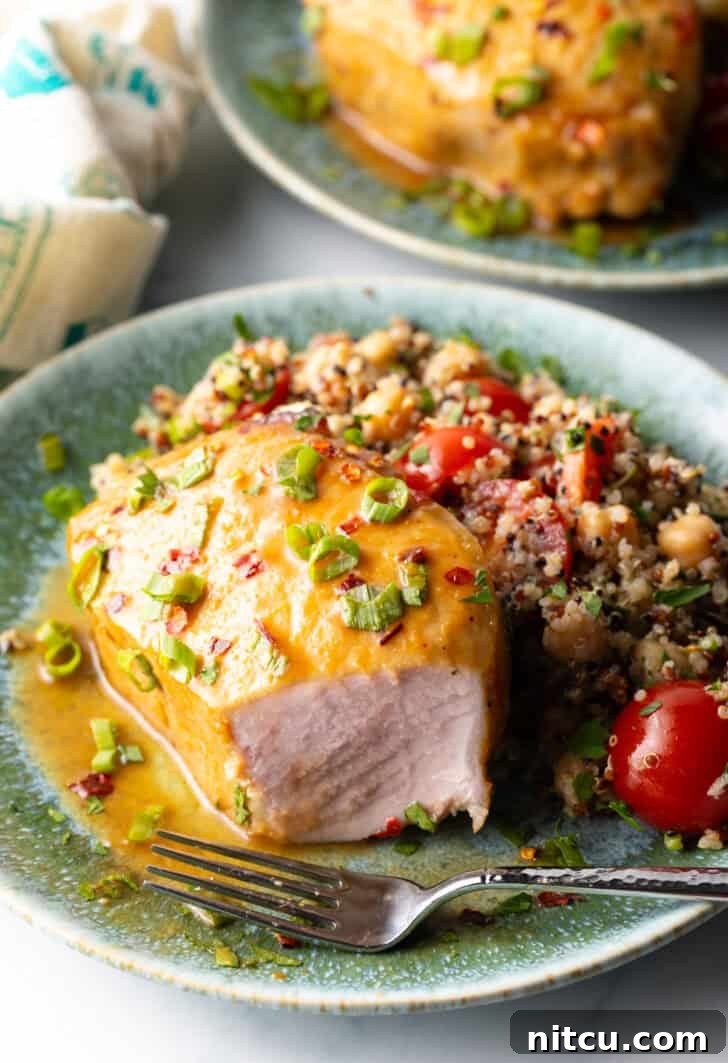 Tender Baked Pork Chops 11 A perfectly baked pork chop, covered with a shiny glaze and fresh chopped scallions, on a plate next to quinoa salad. A fork is strategically placed, and a large portion of the front of the chop has been expertly removed to showcase the tender, juicy interior meat.