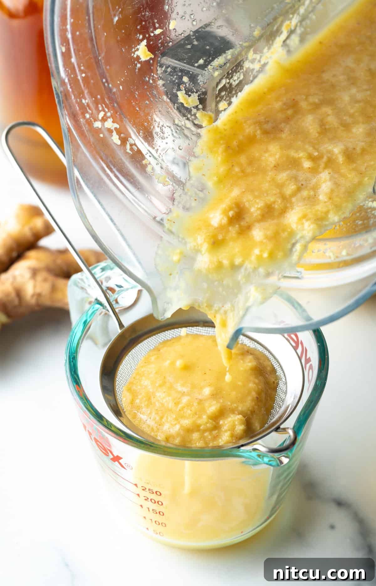 Ginger Zinger Immunity Elixir 5 Pouring pureed ginger mixture from a food processor into a sieve set over a glass measuring pitcher, illustrating the straining process.
