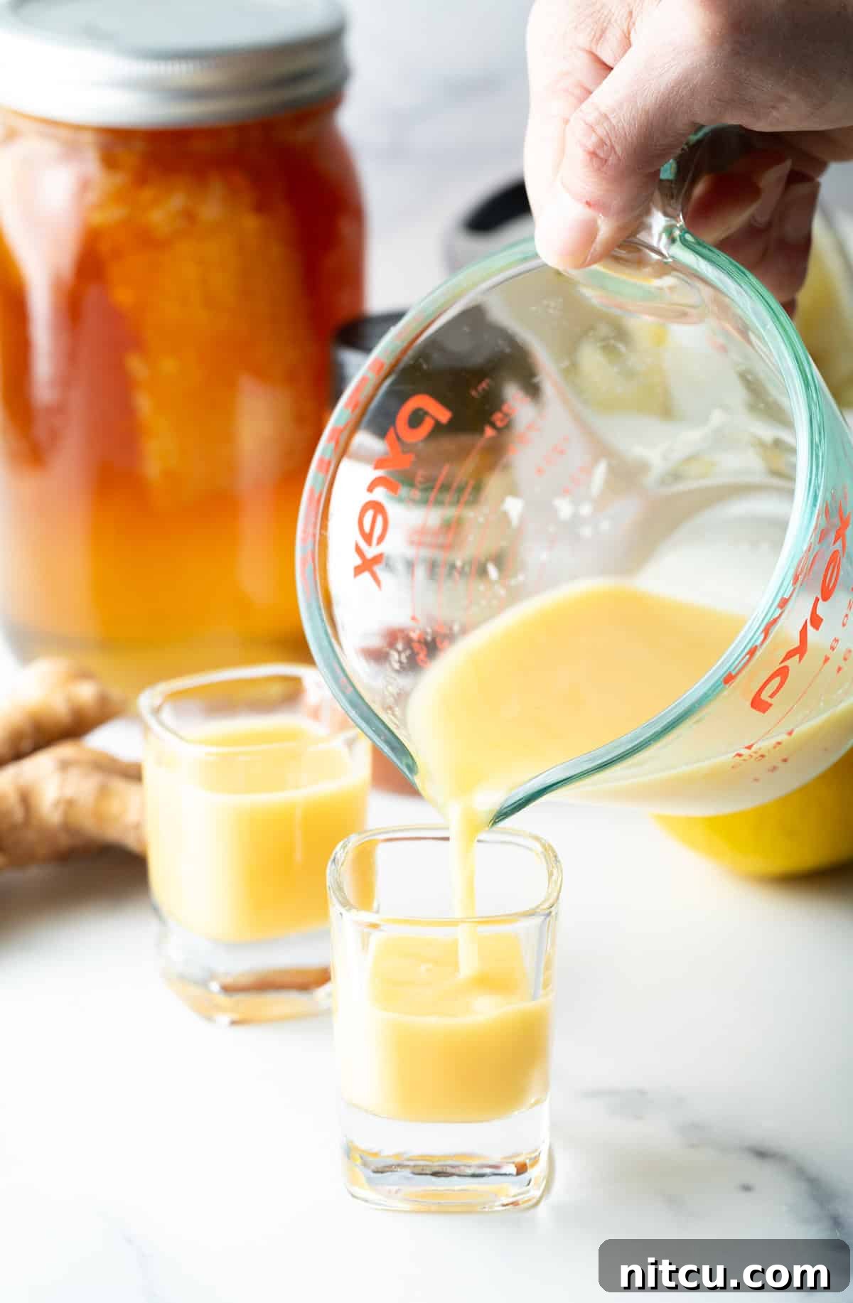 Ginger Zinger Immunity Elixir 6 Pouring vibrant yellow liquid from a glass measuring pitcher into two shot glasses, ready for consumption.