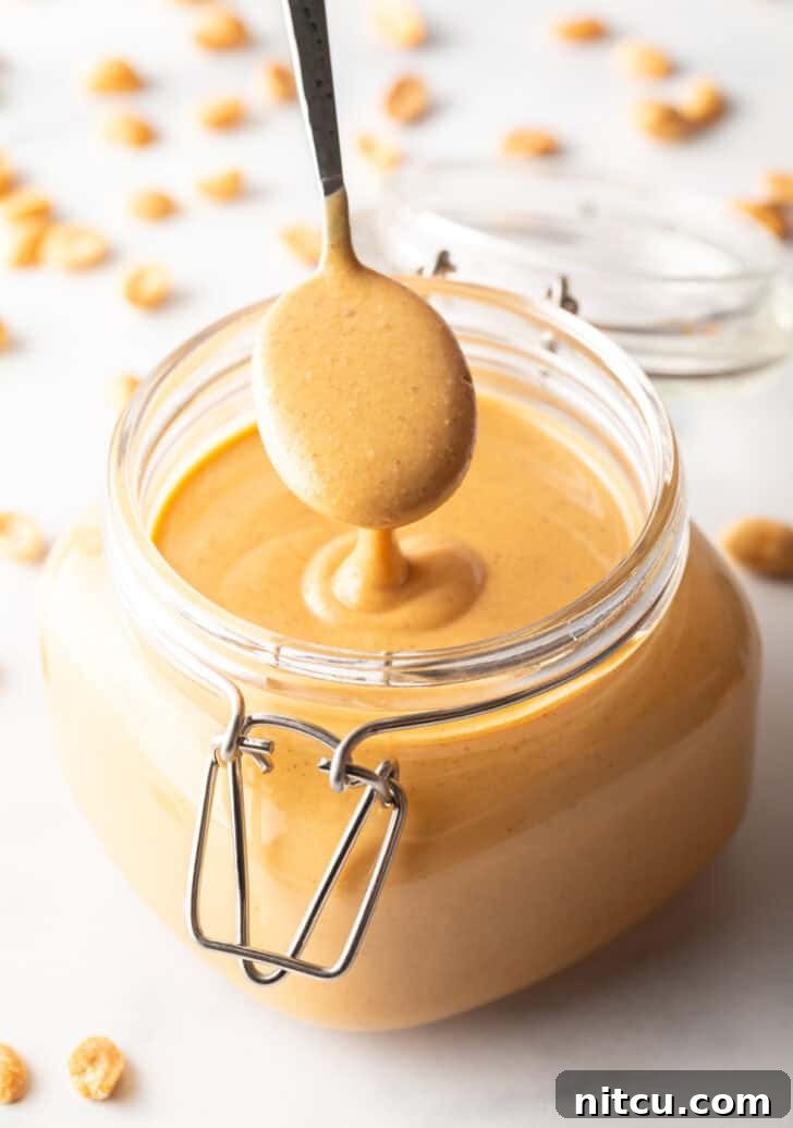 Whip Up Your Own Peanut Butter 2 A spoon taking a scoop of perfectly smooth homemade peanut butter from a clear glass jar, showcasing its creamy texture.