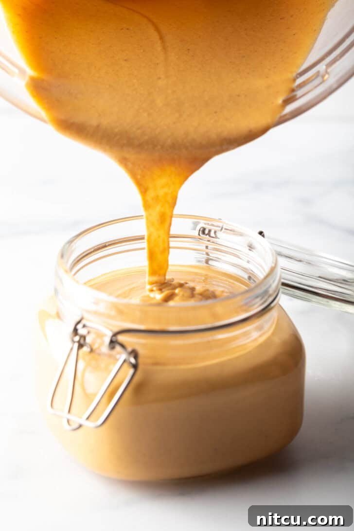 Whip Up Your Own Peanut Butter 11 Smooth homemade peanut butter being poured directly from a blender jar into a clear glass storage jar, highlighting its perfectly liquid consistency.