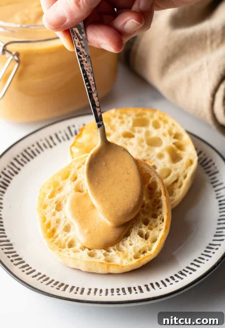 Whip Up Your Own Peanut Butter 12 Hand using a spoon to smooth freshly made homemade nut butter onto half of an English muffin, highlighting its delicious texture.