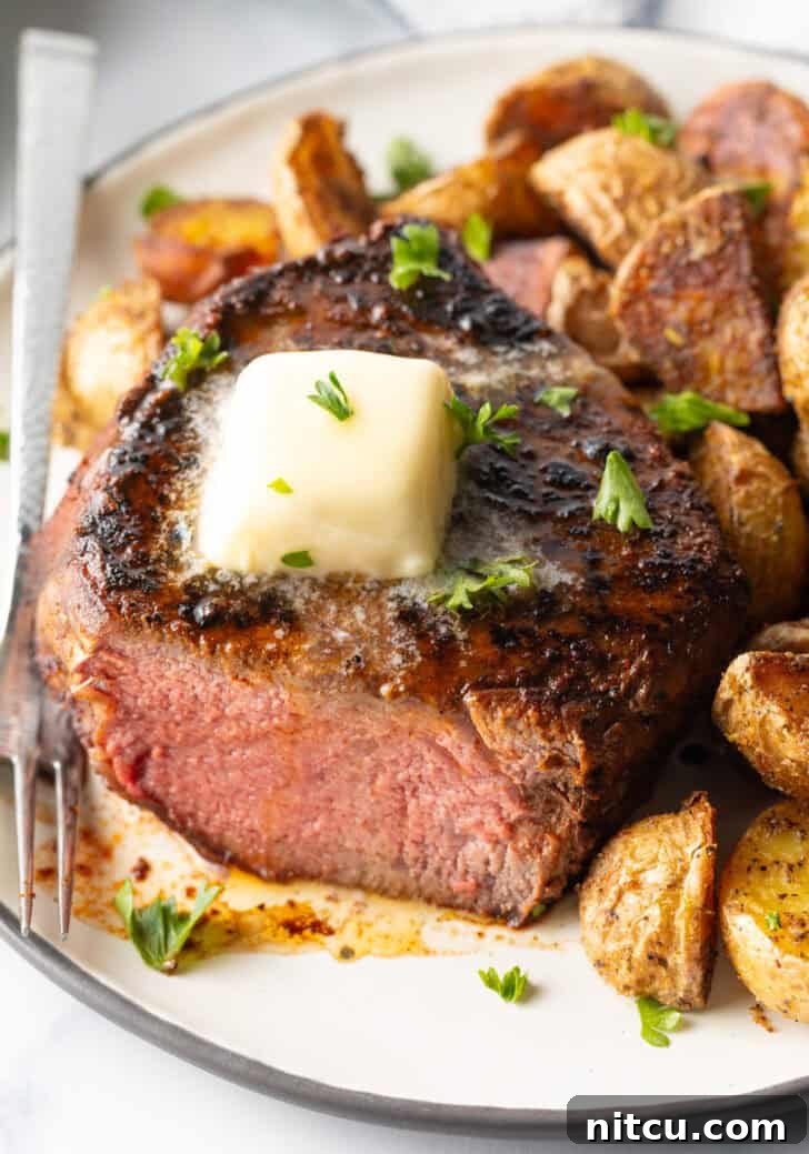 Achieve Steakhouse Quality with Reverse Searing 10 White plate with a reverse sear steak, cut in half to show the medium-rare meat inside. The steak is topped with chopped herbs and a pat of butter. Roasted yellow potato pieces are on the right side of the meat.