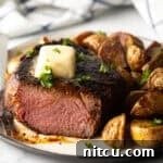 White plate with a reverse sear steak, cut in half to show the medium-rare meat inside. The steak is topped with chopped herbs and a pat of butter. Roasted yellow potato pieces are on the right side of the meat.