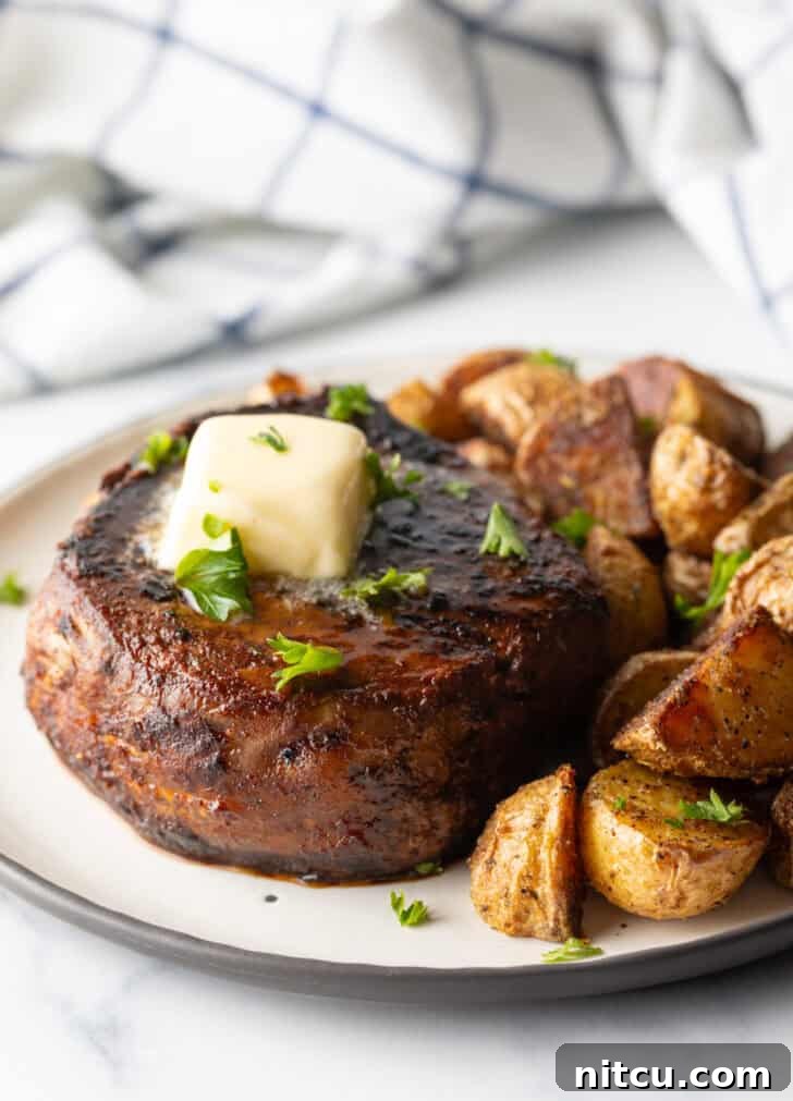 Achieve Steakhouse Quality with Reverse Searing 8 White plate with a reverse sear steak, topped with chopped herbs and a pat of butter. Roasted yellow potato pieces are on the right side of the meat.