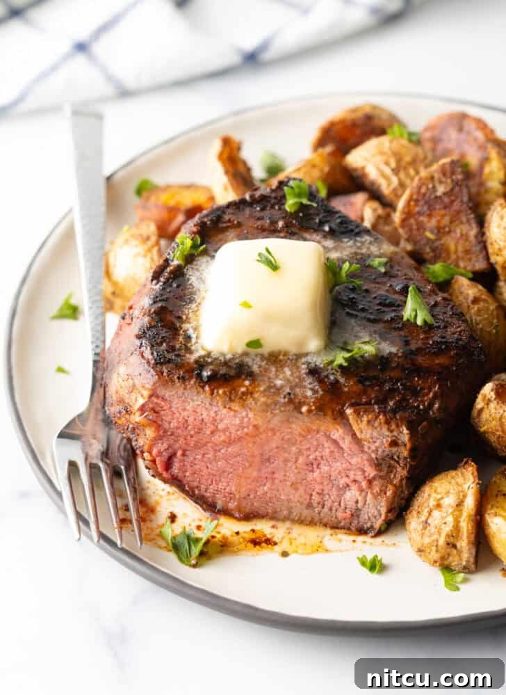 Achieve Steakhouse Quality with Reverse Searing 9 White plate with a reverse sear steak, cut in half to show the medium-rare meat inside. The steak is topped with chopped herbs and a pat of butter. Roasted yellow potato pieces are on the right side of the meat.