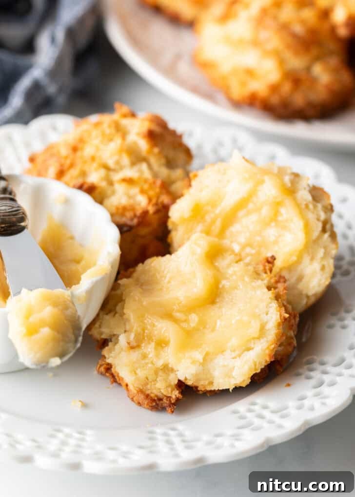 A soft, golden drop biscuit, perfectly split in half on a white plate, with each half generously smeared with glistening whipped honey butter.