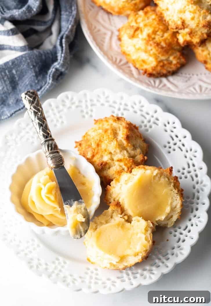 A rustic white plate featuring two golden-brown drop biscuits, accompanied by a small white ramekin of fluffy whipped honey butter with a miniature spreading knife.