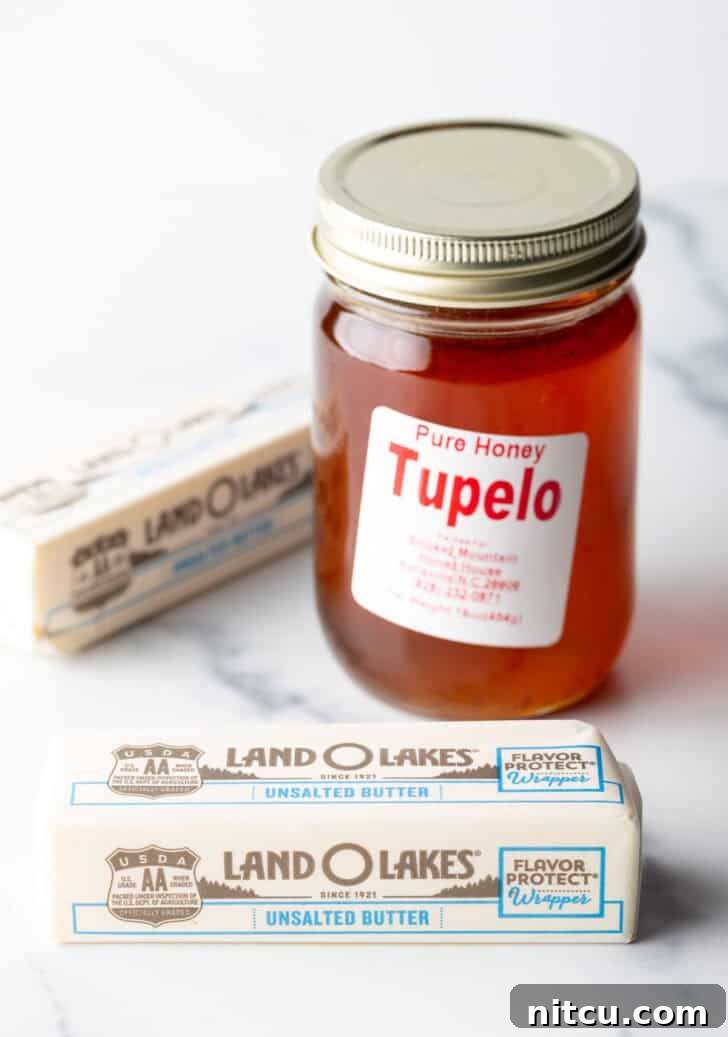 Recipe ingredients: A clear glass jar brimming with golden honey positioned next to two individually wrapped sticks of butter, ready for preparation.