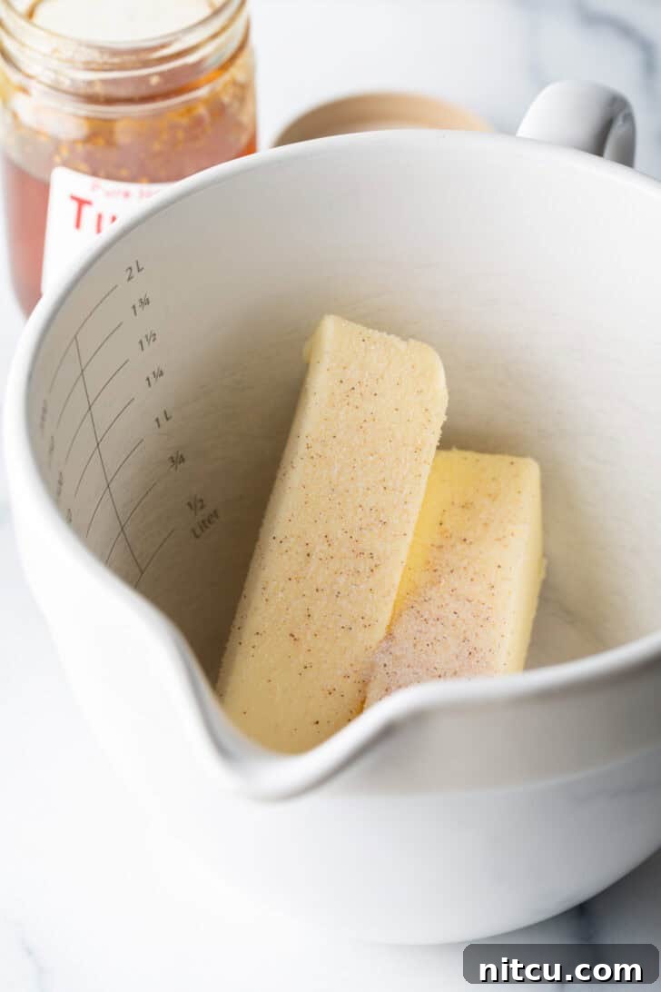 Two sticks of softened butter in a large white mixing bowl, with small piles of various spices arranged around them, suggesting flavor additions.