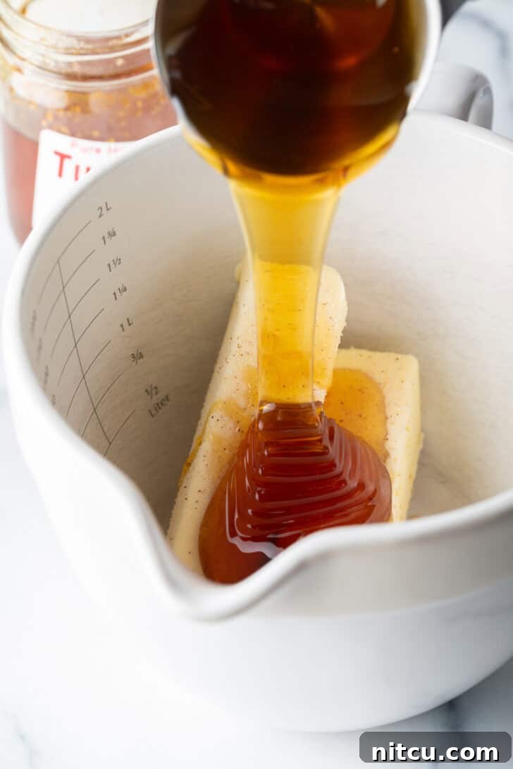 A steady stream of amber honey being poured over two sticks of creamy softened butter within a spacious white mixing bowl, ready for whipping.