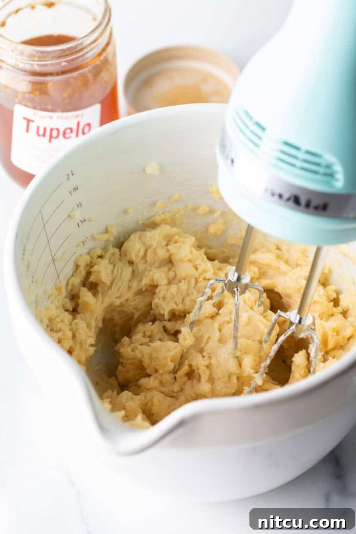 An electric hand mixer actively blending softened butter, golden honey, and a sprinkle of seasonings in a large white mixing bowl, creating a smooth mixture.