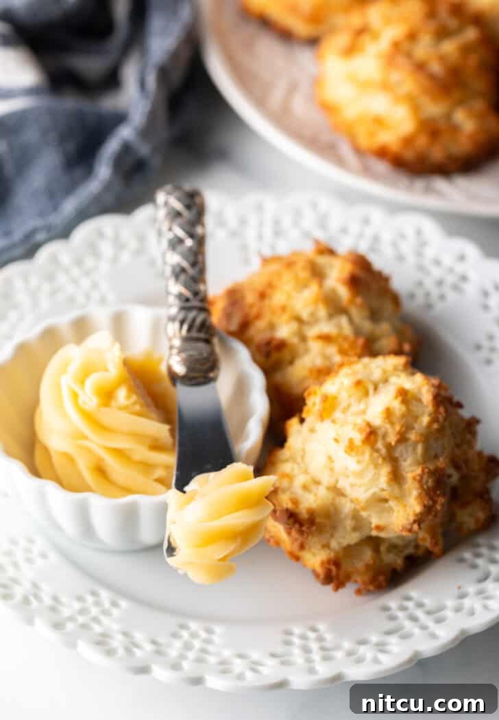 A bright white plate holding two drop biscuits, one split and generously spread with whipped honey butter, alongside a small ramekin of the butter with a spreading knife.