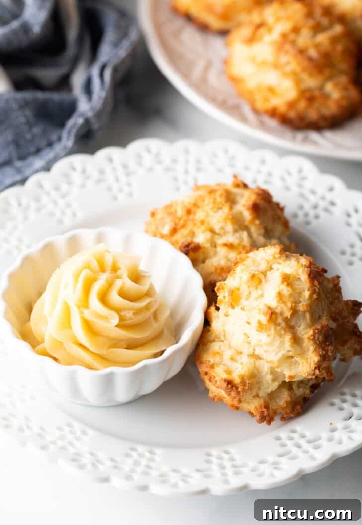 Pillowy Buttermilk Drop Biscuits 12 Two buttermilk drop biscuits on a white plate with a white ramekin of whipped honey butter.