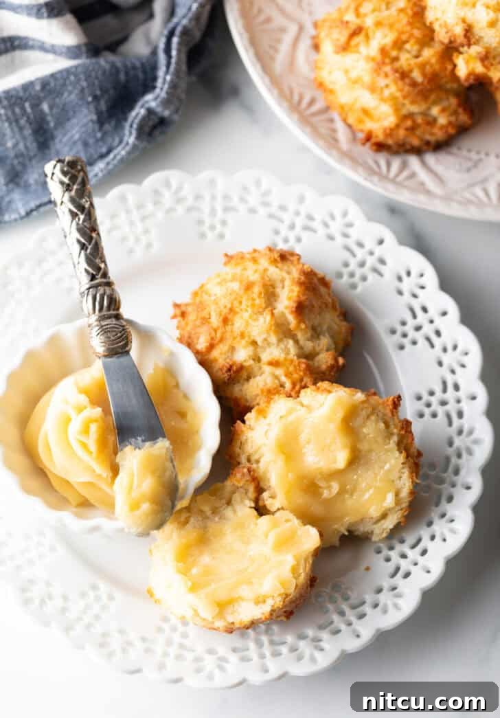 Pillowy Buttermilk Drop Biscuits 4 Close view of two biscuits on a white plate, with a white ramekin of whipped honey butter and a butter knife showing a bit to camera. One biscuit is open with both sides buttered.