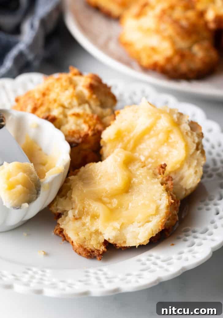 Pillowy Buttermilk Drop Biscuits 5 Close view of two buttermilk drop biscuits on a white plate, with a white ramekin of whipped honey butter and a butter knife showing a bit to camera. One biscuit is open with both sides buttered.