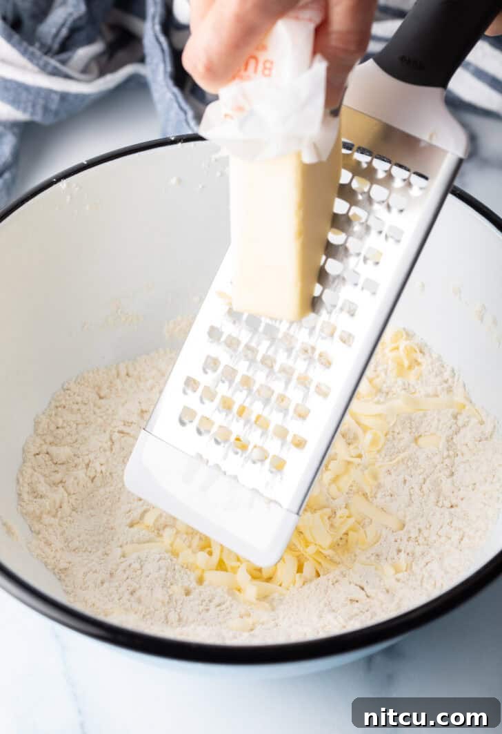 Pillowy Buttermilk Drop Biscuits 7 Using a cheese grater to grate butter into a white bowl with dry biscuit ingredients.