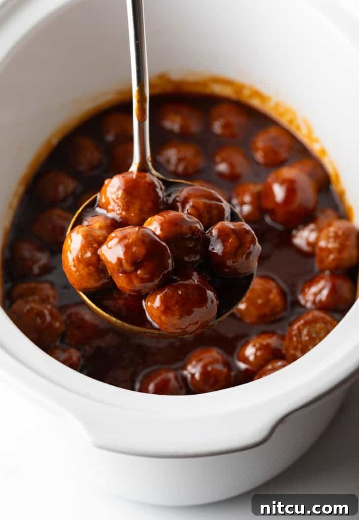 Sweet & Smoky Slow Cooker Meatballs 7 White slow cooker filled with meatballs in bbq sauce. A ladle full of meatballs in being held to camera.