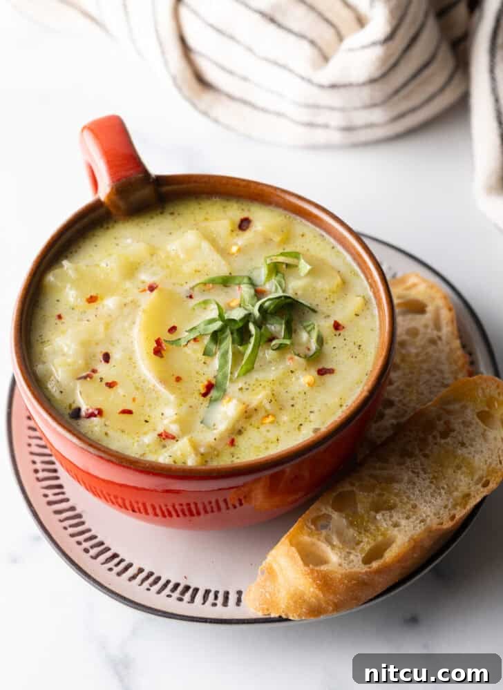 Red oversized mug filled with leek and potato soup, on a plate with slice of baguette on the side.