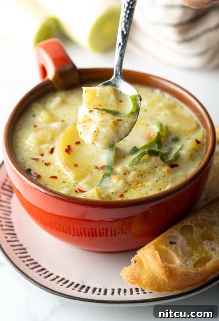 Red oversized mug filled with potato leek soup, on a plate with slice of baguette on the side. A spoon is showing a spoonful of soup to camera.