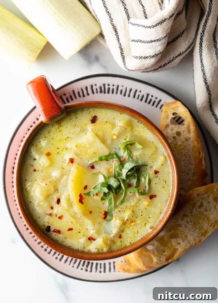 Top down view of an oversized mug filled with potato leek soup, on a plate with slice of baguette on the side.