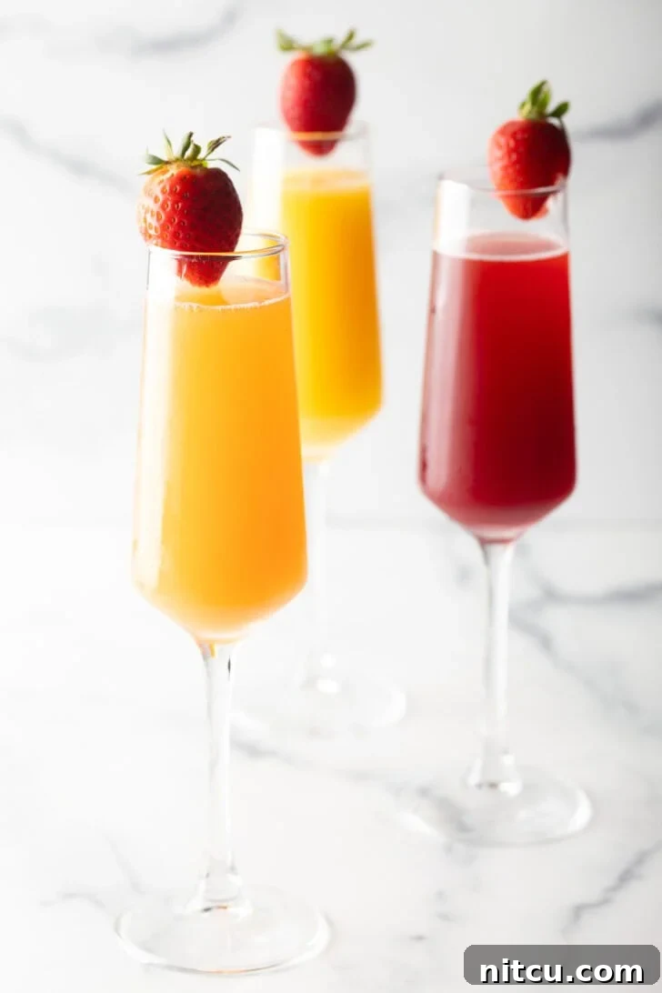 The Ultimate Mimosa Recipe 2 Two champagne flutes filled with orange mimosa, and another glass filled with a cranberry mimosa. Both glasses are garnished with a whole strawberry on the rim.