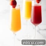 The Ultimate Mimosa Recipe 12 Two champagne flutes filled with orange mimosa, and another glass filled with a cranberry mimosa. Both glasses are garnished with a whole strawberry on the rim.
