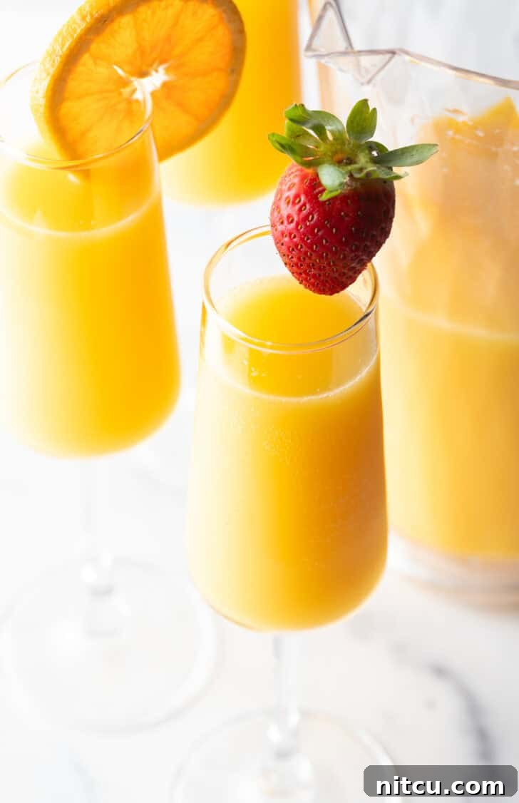 The Ultimate Mimosa Recipe 4 Two glasses of mimosas, one garnished with a large strawberry on the rim, the other garnished with an orange slice on the rim.