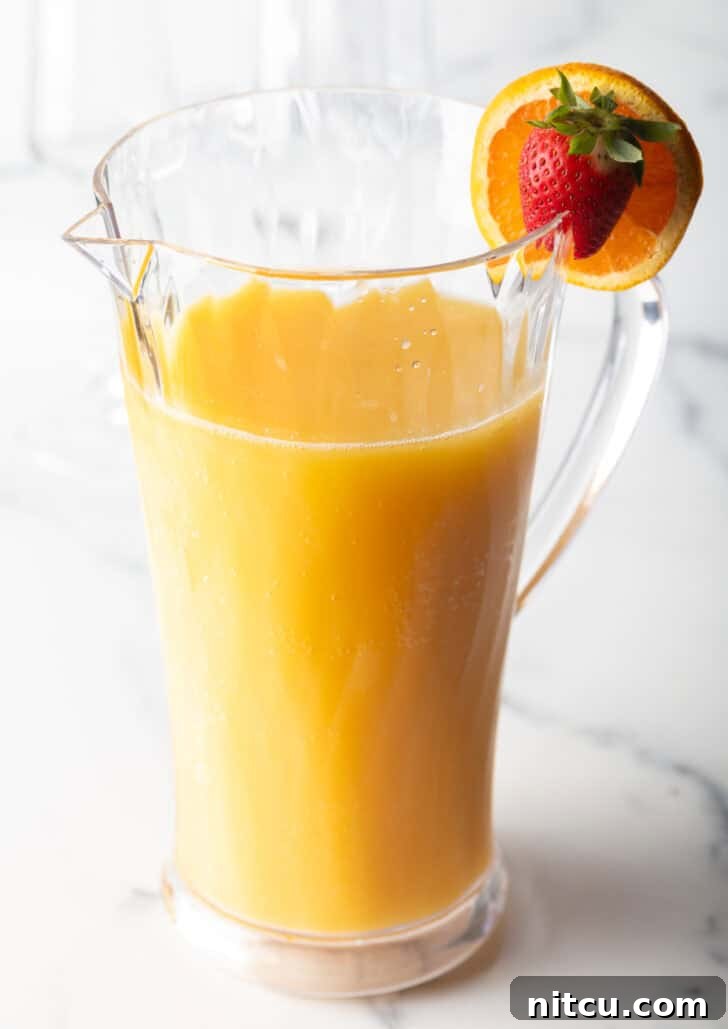 The Ultimate Mimosa Recipe 8 Large glass pitcher of mimosa with an orange and strawberry garnish on the rim.