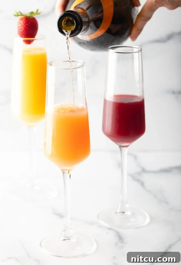 The Ultimate Mimosa Recipe 10 3 champagne glass against a white marble backdrop. The glass in the background is full of orange juice and garnished with a whole strawberry on the rim. In the foreground are a glass half full with cranberry juice, and a glass half full of orange juice with sparkling wine being poured in to the glass.