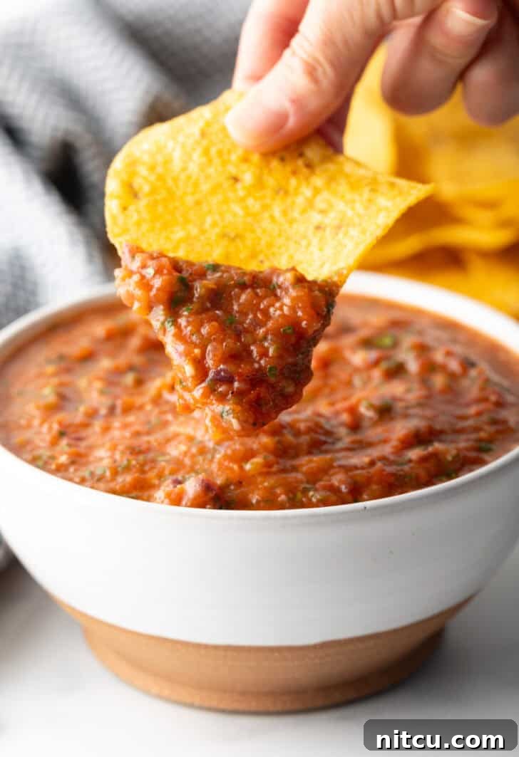 Charred Tomato Salsa 13 White bowl of roasted tomato salsa. A hand is showing a scoop of salsa on a tortilla chip to camera.
